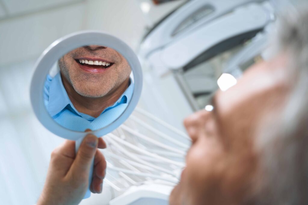 歯科のいすに座り、手鏡を持って口元を確認する高齢男性
