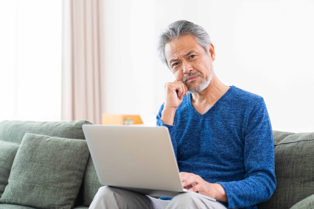 ソファに座り、パソコンを膝に乗せて考え事をしている高齢男性